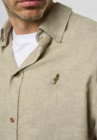 Beige linnen overhemd met een kraag, voorzien van bruin knopen en een klein groen geborduurd detail op de borst. Glad, lichtgewicht textuur.