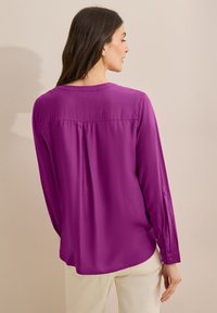 Blusa de manga larga en un vibrante color púrpura, hecha de una suave tela. Presenta una espalda plisada, puños abotonados y un escote redondeado.