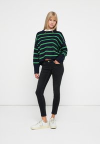 Mujer con un suéter a rayas en tonos azul marino y verde, jeans ajustados negros y zapatillas blancas, de pie frente a un fondo blanco liso.