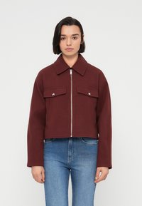 Μπορντό cropped μπουφάν από απαλό υλικό, με φερμουάρ μπροστά, δύο τσέπες στο στήθος και κλασικό γιακά. Συνδυάζεται με μπλε τζιν.