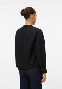 Svart blus med hög krage och långa ärmar, prydd med veck på baksidan och guldknapp-accenter på manschetterna. Tyget ser slätt ut.