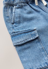 Nærbillede af lyseblå denim bukser med sidelomme og klaplomme med synlige syninger samt elastisk taljebånd med hvid snor.
