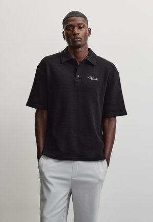 Homme portant un polo noir texturé à manches courtes avec un logo en écriture blanche et un pantalon gris clair, debout devant un fond clair uni.