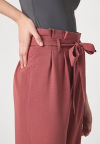 Rose-colored, high-waisted trousers with a tied waist, side pockets, and visible pleats. Fabric has a textured appearance.