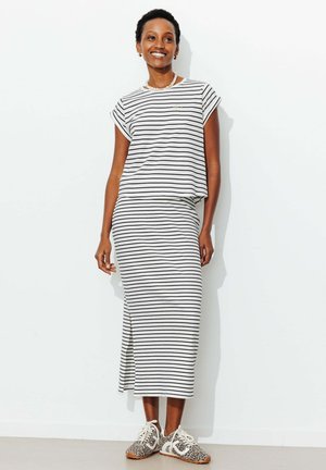 Femme souriante portant une robe blanche à rayures horizontales noires et des baskets à imprimé léopard, debout devant un mur blanc uni.