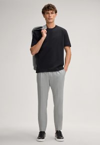 Zwart T-shirt met korte mouwen, lichtgrijze op maat gemaakte broek met subtiele textuur, en zwarte sneakers, met een grijze jas over de rechterarm gedrapeerd.