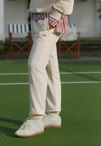 Kremowe dresowe spodnie o dopasowanym kroju, z czerwonym haftem "self.", zestawione z białymi trampkami z gumową podeszwą. Tło z trawą.