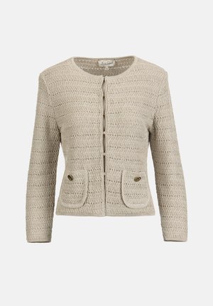 Žena v béžovom štruktúrovanom zapínanom saku a zodpovedajúcej sukni po kolená, držiaca krémovú kabelku so zlatým reťazcom, stojaca na bielom pozadí.