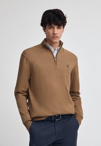 Brauner Reißverschluss-Pullover aus weichem Stoff, mit geripptem Kragen und Bündchen. Gesticktes Logo auf der linken Brust. Getragen mit dunklen Hosen.