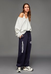 Ljusgrå oversize tröja med v-ringning, kombinerad med mörkblåa mjukisbyxor med vit text. Avslutas med silverfärgade sneakers.