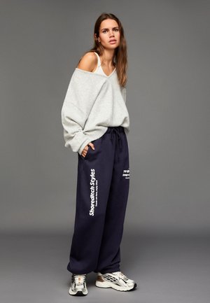 Svetlošedý oversized sveter s výstrihom do V, skombinovaný s tmavomodrými teplákmi s bielym nápisom. Dokončené striebornými teniskami.