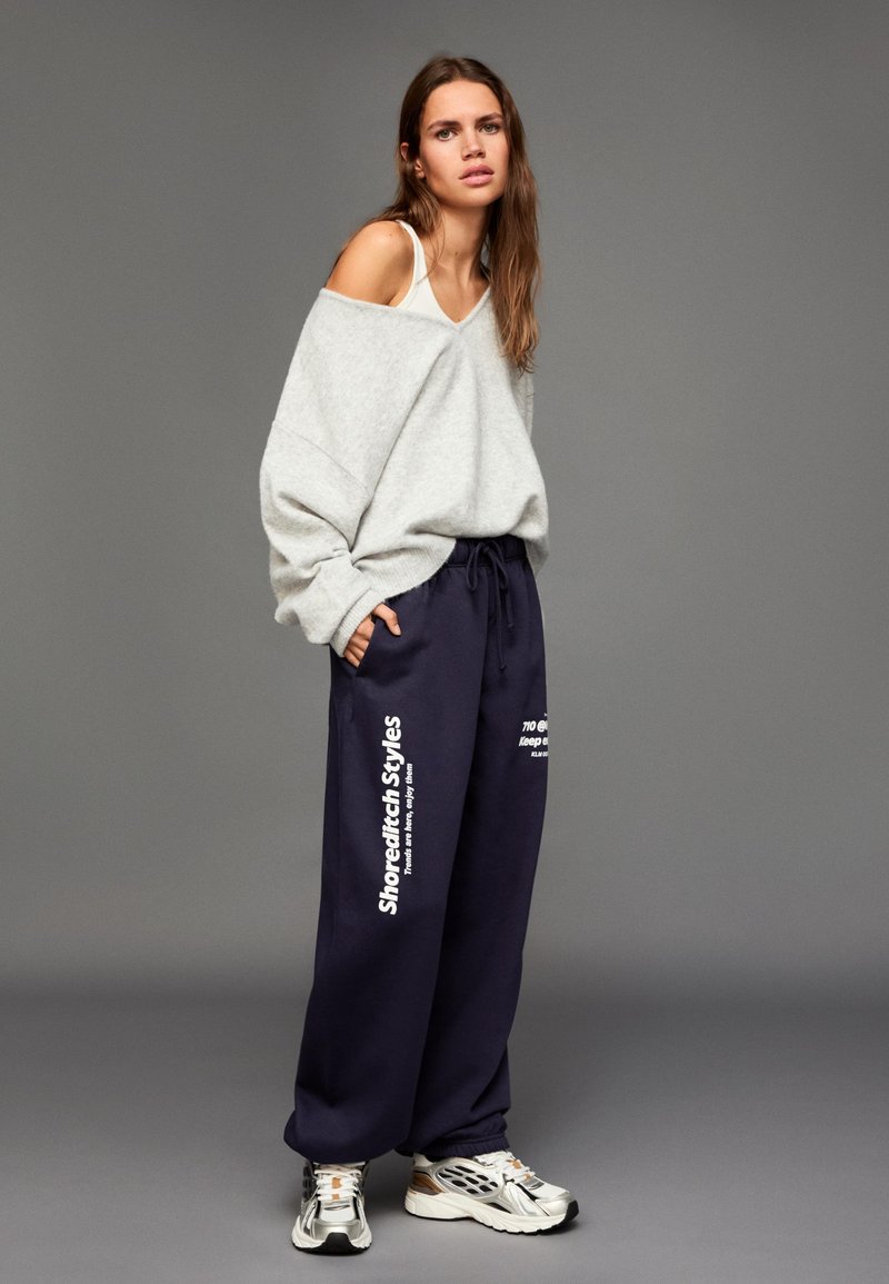 Ljusgrå oversize tröja med v-ringning, kombinerad med mörkblåa mjukisbyxor med vit text. Avslutas med silverfärgade sneakers.