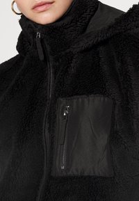 Czarna polarowa kurtka z wysokim kołnierzem, wyposażona w gładką kieszeń na zamek na lewej piersi oraz miękkie, teksturowane body.