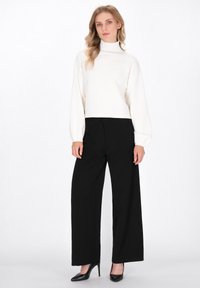 Vrouw met golvend blond haar draagt een witte coltrui, zwarte wijde pantalon en zwarte hoge hakken, staande voor een witte achtergrond.