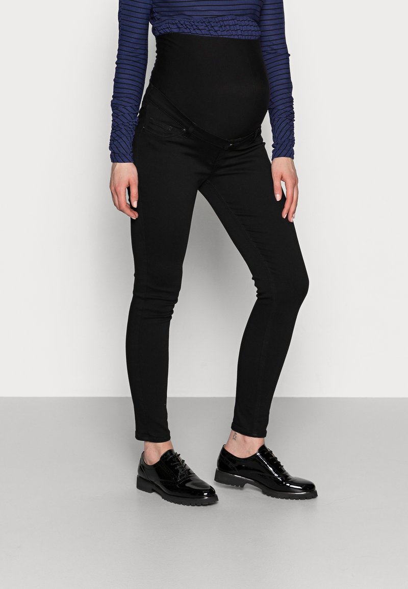 Personne enceinte portant un pantalon de maternité noir, un haut à manches longues bleu rayé, et des chaussures Oxford noires brillantes, debout sur un sol gris.
