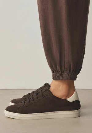 Brown suede low-top sneakers with white heel detail worn with brown cuffed pants on a beige floor.