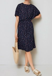 Námořnické šaty s krátkými rukávy, květinovým potiskem, v pase stažené a délky ke kolenům. Spárované se zlatými teniskami a béžovou tkanou taškou.