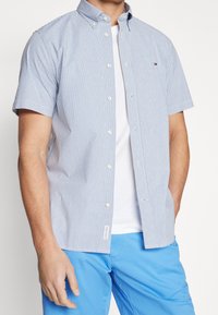 Camicia a maniche corte con bottoni di colore azzurro chiaro, caratterizzata da strisce verticali bianche, colletto classico e una patch con logo sul petto. Abbinata a pantaloni corti blu.