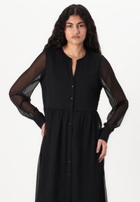 Μαύρο φόρεμα με κουμπιά και μακριά διαφανή μανίκια, συγκεντρωμένη μέση και ίσια γραμμή. Διαθέτει γιακά και λειτουργικό κούμπωμα με κουμπιά.