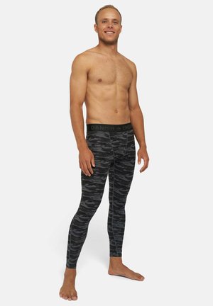 Hombre sonriente sin camisa, de pie descalzo, vistiendo leggings de patrón de camuflaje en negro y gris con cinturilla de marca sobre fondo blanco.