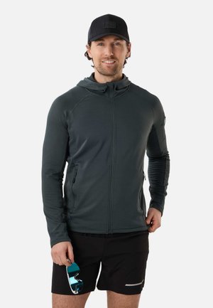 Mann trägt dunklen Hoodie mit Reißverschluss, schwarze Kappe und schwarze Shorts, hält blaue reflektierende Sonnenbrille in der rechten Hand und lächelt in die Kamera.