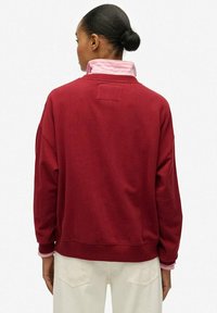 Sudadera de algodón roja con un corte holgado, que presenta un alto cuello y un cuello a rayas rosa en capas por debajo. Diseño sencillo, puños acanalados.