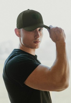 Gorra verde oliva con un emblema de concha texturizada, que presenta un visera curva. La lleva una persona con una camisa negra de manga corta, que muestra un brazo musculoso.