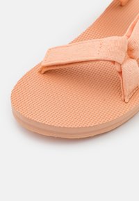 Chancla de color melocotón con suela de goma texturizada y tira de tela. Material suave, diseño plano, con un sutil patrón en la plantilla.