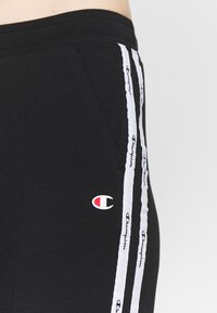 Černé sportovní kalhoty s bílými bočními pruhy a opakovaným textem. Obsahuje malé logo v červené a bílé barvě. Měkká, elastická látka s kapsami.