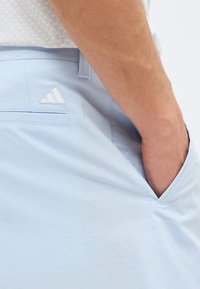 Κοντινό πλάνο του χεριού ενός ατόμου στην εμπρόσθια τσέπη ανοιχτόχρωμων μπλε σορτς με ένα μικρό λευκό κεντημένο λογότυπο κοντά στην πίσω τσέπη.