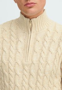 Beige Kabelstrickpullover mit hohem geripptem Kragen und halbem Reißverschluss. Verfügt über strukturierte Muster und einen weichen, gewebten Stoff.