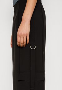Pantalon noir ajusté avec une bande latérale ornée d'un anneau argenté. Le tissu est lisse, conçu pour une silhouette cintrée.