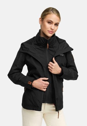 Femme portant une veste noire zippée sur un ensemble beige, ajustant le col de la veste, avec une expression faciale calme sur un fond uni.