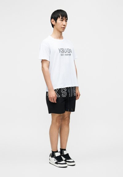 Λευκό βαμβακερό t-shirt με τυπωμένο το "KSUBI EST. 1999" μπροστά, σε συνδυασμό με μαύρο σορτς με μεγάλα λευκά γράμματα. Μαύρα αθλητικά παπούτσια με λευκές λεπτομέρειες.