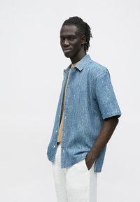 Rövid ujjú, galléros ing világoskék farmerből; laza szabású, látható varrással és fehér gombos záródásokkal elöl. Bézs felső felett viselve.