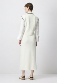 Witte gebreide vest over een wit shirt, gecombineerd met een bijpassende lange rok. Heeft een geribde zoom en contrasterende zwarte accenten. Casual schoeisel.