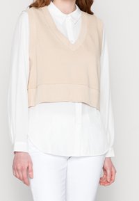 Chaleco corto beige con escote en V, textura acanalada, superpuesto a una camisa blanca con botones y mangas largas, combinado con pantalones blancos.