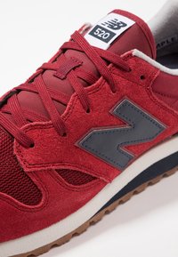 Detailní záběr na červen�é semišové a síťované tenisky New Balance 520 s červenými tkaničkami, černým logem "N" a bílou mezipodešví na světlém pozadí.