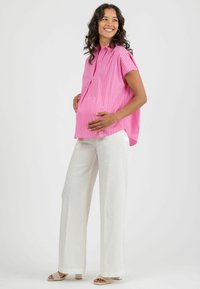Blusa a righe rosa con maniche corte, vestibilità rilassata, abbinata a pantaloni bianchi a gamba larga. Il modello sta in piedi con una mano sul pancione.