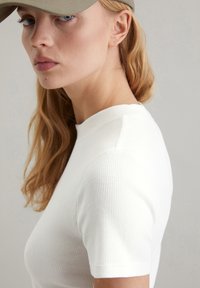 Weißes, geripptes Baumwoll-T-Shirt mit Rundhalsausschnitt, getragen mit einer geschlossenen olivgrünen Cap. Glatte Textur, taillierte Passform und kurze Ärmel.