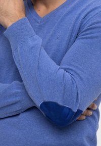 Homme aux bras croisés portant un pull bleu col en V avec des coudières bleu foncé et un petit logo brodé sur la poitrine.