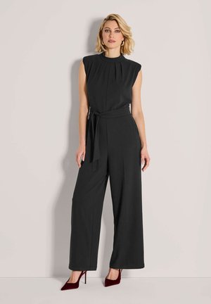 Frau steht in einem schwarzen ärmellosen Jumpsuit mit weitem Bein und Taillengürtel, trägt burgunderfarbene High Heels vor einem beigen Hintergrund.