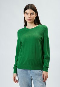 Grüner gestrickter Pullover mit geripptem Rundhalsausschnitt, strukturiertem Obermuster und langen Ärmeln. Kombiniert mit hellblauen Jeans.