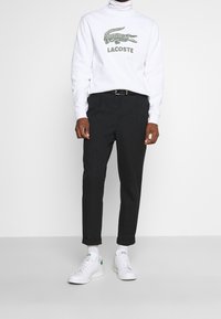 Sudadera de cuello alto blanca con logo verde de Lacoste, combinada con pantalones negros y zapatillas blancas con acentos verdes.