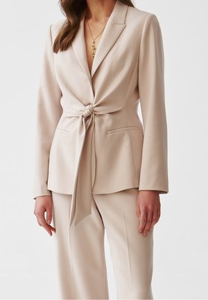 Kobieta ubrana w beżowy, dopasowany garnitur z długimi rękawami, marynarkę z wiązanym węzłem oraz pasujące, wysokie spodnie na tle jednolitego tła.