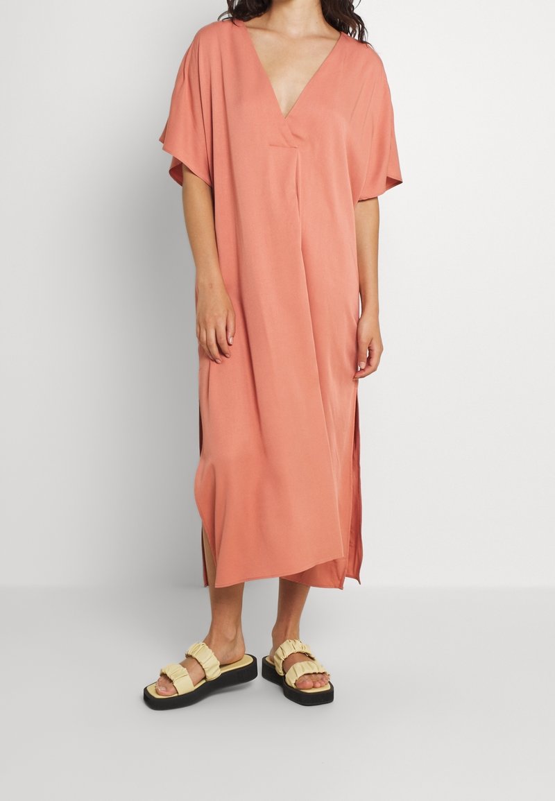 Pfirsichfarbiges, knielanges Kleid mit V-Ausschnitt, kurzen Ärmeln und seitlichen Schlitzen. Kombiniert mit cremefarbenen Plateau-Sandalen mit Rüschenakzenten.