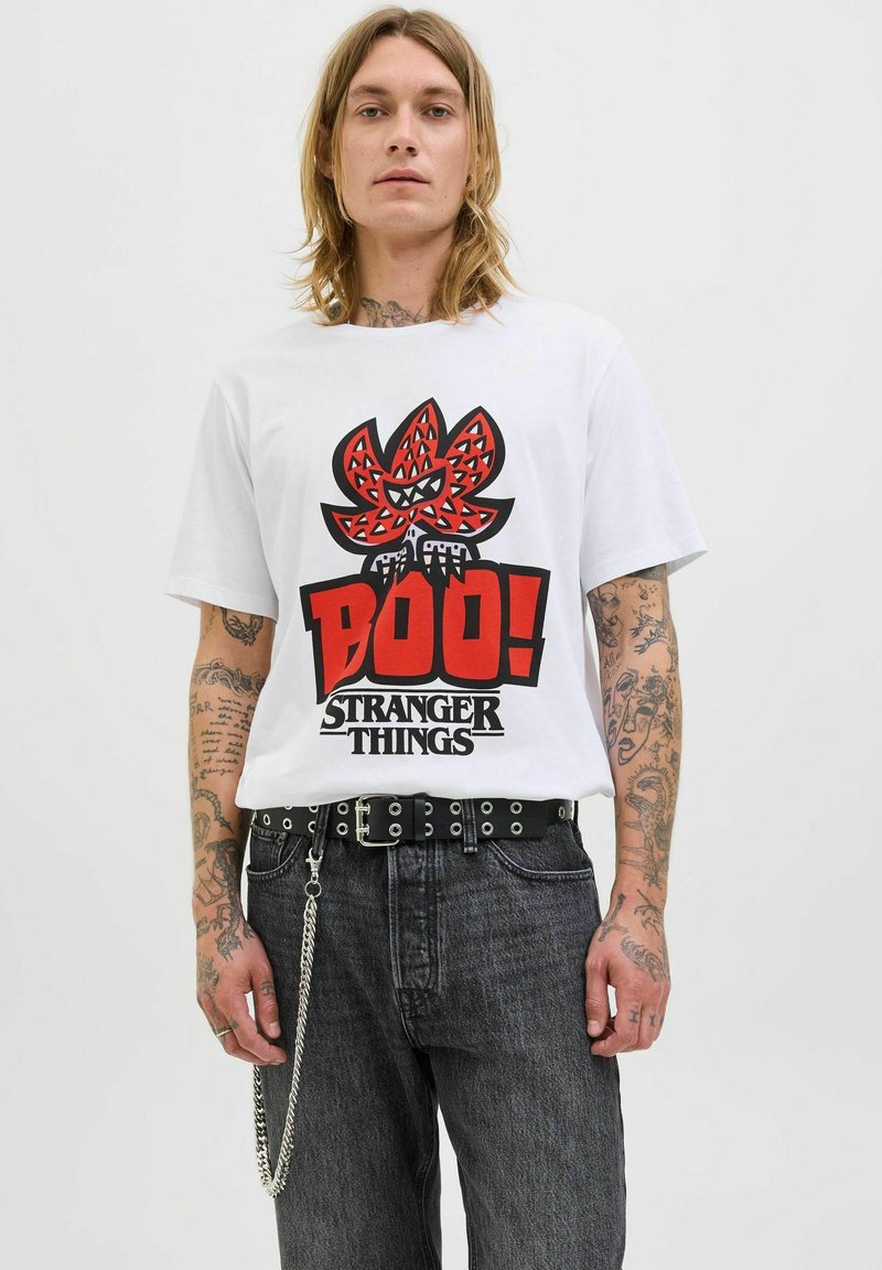 Tricou alb din bumbac cu un grafic îndrăzneț roșu și negru, având textul "Boo!" și "Stranger Things". Asortat cu blugi închisi la culoare și o curea neagră.