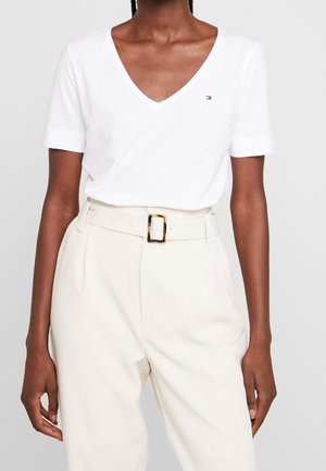 Vrouw draagt een wit V-hals top met korte mouwen en een klein logo, gecombineerd met crème broek met hoge taille, brede riem en schildpadkunststof gesp.