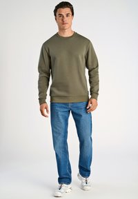 Sudadera de manga larga verde oliva con puños y dobladillo acanalados, combinada con jeans de color azul claro y zapatillas deportivas blancas.