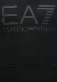 Čierna látka s potlačeným tmavosivým logom EA7 Emporio Armani, ktoré obsahuje štýlizované „EA7“ a emblém orla medzi slovami.